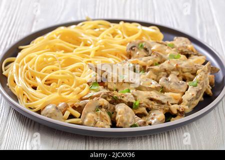 Kälber Leberstroganoff mit Rahm, Vollkornsenf, Zwiebeln und Pilzen, serviert mit Spaghetti auf dem Teller auf dem weißen Holztisch, horizontale Ansicht Stockfoto