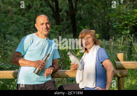 Reifes Paar, das nach dem Training im Park mit wiederverwendbaren Metallflaschen zur Kamera schaut Stockfoto