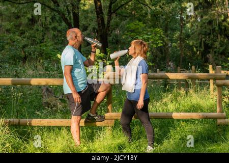 Seitenansicht des Trinkwassers eines älteren Mannes und seiner Frau aus einer wiederverwendbaren, umweltfreundlichen Flasche im Park Stockfoto