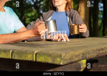 Nicht erkennbares Seniorenpaar hält und trinkt aus Metallbechern auf einem Campingtisch, während es redet Stockfoto