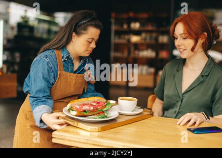 Kellnerin mit Down-Syndrom serviert einem Gast ein Sandwich und Kaffee in einem trendigen Café. Professionelle Frau mit einer intellektuellen Behinderung, die in einem Stockfoto