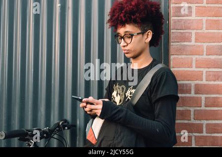 Junge lateinamerikanische Brünette Radfahrerin, mit Afro, Brille getragen und in schwarz gekleidet, stehend auf der Straße, die Kartenroute am Telefon zu überprüfen, mit Stockfoto