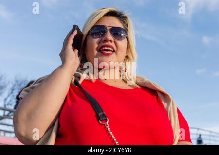 Porträt schöne junge lateinische kaukasische plus size-Modell Frau im Freien reden am Telefon mit Sonnenbrille und roter Kleidung, bei Sonnenuntergang in der buenos Stockfoto