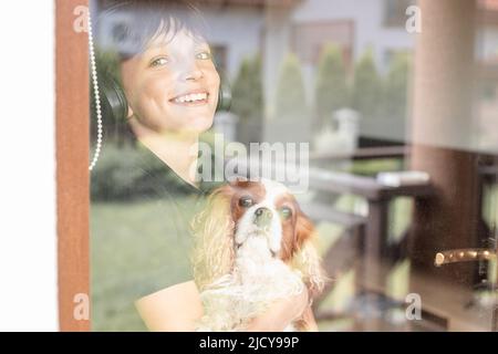 Porträt eines lächelnden fröhlichen Teenagers Junge mit Hund Cavalier King Charles spaniel, durch das Fenster schauen, posieren. Stockfoto