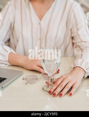 Nahaufnahme einer nicht erkennbaren Frau, die an einem Tisch mit Laptop, einem Glas Wasser und Gläsern sitzt Stockfoto