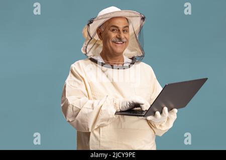 Reifer männlicher Bienenhalter in Uniform mit einem Laptop, der auf blauem Hintergrund isoliert ist Stockfoto