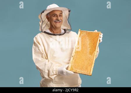 Reifer männlicher Bienenhalter in einer Uniform, der einen Honigbienenrahmen auf blauem Hintergrund isoliert hält Stockfoto