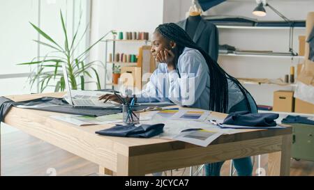 Ein talentierter afroamerikanischer Modedesigner kreiert eine neue Modekollektion, während er in einem Fashion Business Startup Studio an einem Laptop arbeitet. Konzept Stockfoto