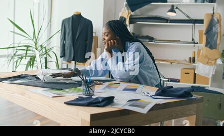 Ein talentierter afroamerikanischer Modedesigner kreiert eine neue Modekollektion, während er in einem Fashion Business Startup Studio an einem Laptop arbeitet. Konzept Stockfoto