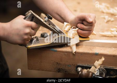 Nahaufnahme von abgeschnittenen Tischlerhänden, die mit einem Plane Equipment Tool auf der Werkbank arbeiten. Zimmerei Hobel in der Werkstatt Stockfoto