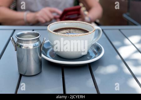 Eine Frau ist damit beschäftigt, SMS auf ihrem Handy zu schreiben, während sie auf eine frisch zubereitete Tasse Kaffee wartet, um abzukühlen. An einem Sommerabend sitzt sie an einem Kaffeetisch im Freien Stockfoto