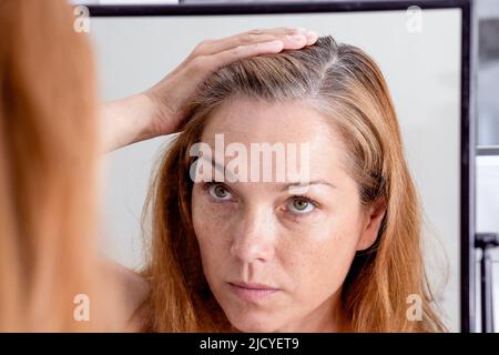 Grauhaarige kaukasische Frau mittleren Alters, die in Spiegelreflexion den grauen Haarkopf anschaut Stockfoto