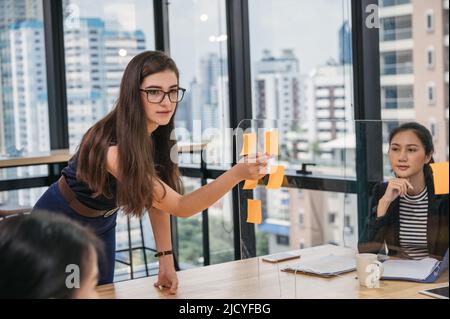 Die junge kaukasische Geschäftsfrau klebt eine Haftnotiz auf die Trennwand und diskutiert während des Treffens mit einem Mitarbeiter im modernen Büro über Geschäftsidee Stockfoto