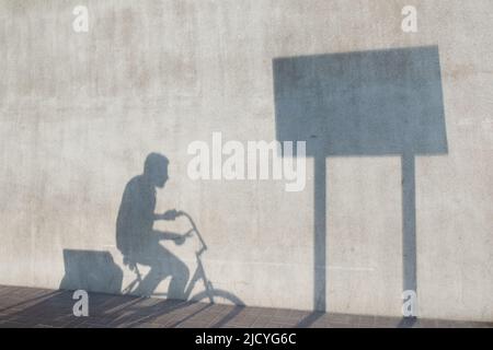 Schatten eines Mannes auf dem Fahrrad, der durch den Schatten eines rechteckigen Straßenschildes auf einer leeren Betonwand fährt. Für konzeptuelle Anzeigen und horizontalen Kopierbereich. Stockfoto