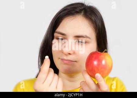 Eine Frau in Gelb hält einen Apfel und eine Pille in ihren Händen. Vitamine in natürlicher Form und in synthetischen Nahrungsergänzungsmitteln Stockfoto