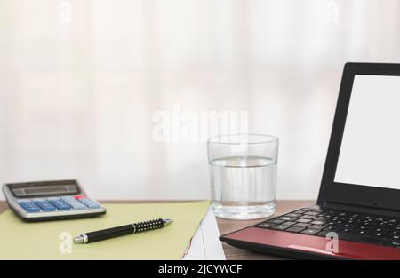 Auf einem Holztisch befinden sich ein Taschenrechner, ein Ordner, ein Stift, ein Laptop und ein Glas Wasser. Stockfoto