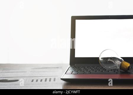 Auf einem Holztisch befindet sich ein Laptop, auf dessen Tastatur sich eine Glühbirne befindet, daneben eine Stromrechnung. Der Computerbildschirm ist leer. Stockfoto
