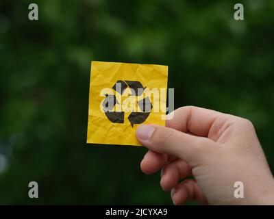 Eine Frau, die eine gelbe Haftnotiz mit einem Recycling-Symbol in der Hand vor einem Hintergrund aus Laub hält. Nahaufnahme. Stockfoto