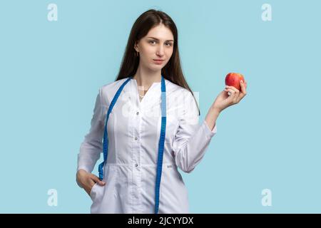 Frau Doktor Ernährungsberaterin trägt weißen Mantel. Gut aussehende Ernährungswissenschaftlerin in medizinischer Uniform mit grünem Apfel und Messband auf blauem Hintergrund Studio. Stockfoto