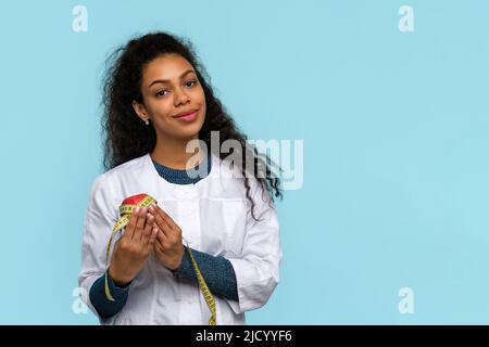 Dunkelhäutige Frau Doktor Ernährungsberaterin trägt weißen Mantel. Gut aussehender Ernährungsberater in medizinischer Uniform mit rotem Apfel und Messband auf blauem Studio Wa Stockfoto