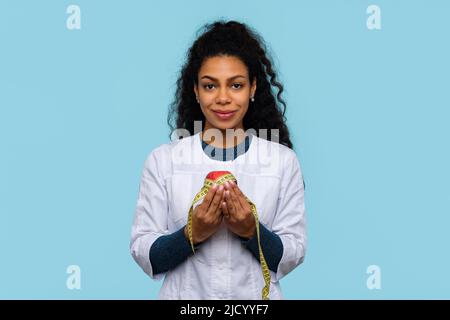 Nicht-kaukasische Hautärztin, Ernährungswissenschaftlerin, trägt White Coat. Gut aussehender Ernährungsberater in medizinischer Uniform mit rotem Apfel und Messband auf Blau Stockfoto