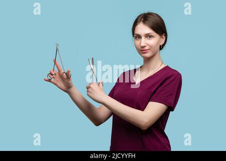 Junge Chirurgin posiert mit Steel Surgical Instruments auf blauem Hintergrund Studio. Ärztin Ärztliche Mitarbeiterin trägt medizinische Uniform Chirurgische Su Stockfoto