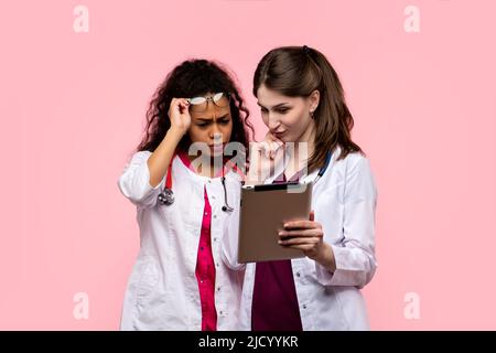 Zwei unterschiedliche Ethnien Medizinstudenten schauen sich Digital Tablet auf Pink Studio Hintergrund an. Praktikanten verwendeten App zur Aufzeichnung der Krankengeschichte, Check Laborato Stockfoto