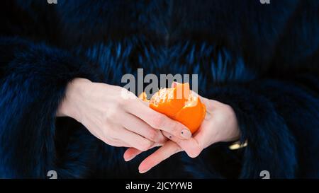 Geschälte Orange Mandarine in Mädchen Hand auf dunkelblauem Pelz Hintergrund im Freien. Nahaufnahme einer gesichtslosen Vegetarierin. Stockfoto