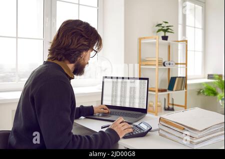 Männlicher Buchhalter, der im Büro sitzt, mit Tabellenkalkulationen auf dem Laptop arbeitet und Rechner benutzt Stockfoto