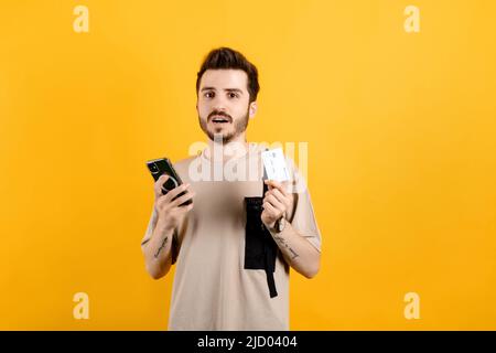 Kaukasischer Mann trägt beiges T-Shirt und posiert isoliert auf gelbem Hintergrund mit Kreditkarte und Smartphone mit enttäuschter Gesichtsausdruck, Gezänk, Konkurs Stockfoto