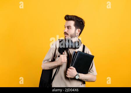 Kaukasischer junger Mann, der T-Shirt trägt und isoliert auf gelbem Hintergrund posiert und mit Rucksack zur Seite blickt. Stockfoto