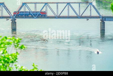 Eine Eisenbahnbrücke, auf der eine Lokomotive fährt, ein Zug über einen Fluss, auf dem ein Motorkautschukboot schwimmt Stockfoto