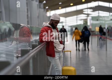 Afroamerikanischer junger Mann mit Smartphone, der im Flughafenterminal mit Gepäck unterwegs ist Stockfoto