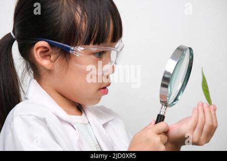 Porträt eines kleinen Mädchens in einer Brille mit einer Lupe und Blick auf Blätter in Forscher- oder Wissenschaftsuniform auf weißem Hintergrund. Wenig Wissenschaft Stockfoto