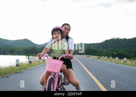 Nettes kleines Mädchen, das mit ihrer Mutter bei Sonnenuntergang auf einer Seestraße ein Fahrrad reitet. Glückliche Familie, die gemeinsam Outdoor-Aktivitäten unternimmt. Gesunde Sommeraktivitäten Stockfoto