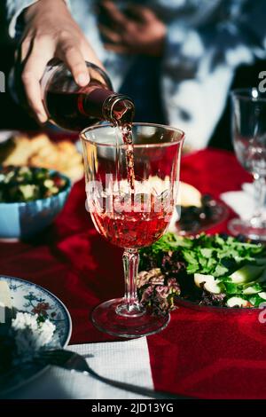 Eine Frau gießt Rotwein in ein Glas auf dem Tisch. Abendessen mit einer großen Gruppe von Freunden auf dem Land. Vertikales Foto. Stockfoto