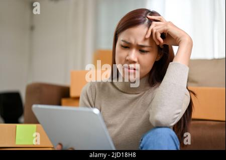 Gestresste junge asiatische weibliche Online-Shopping-Shop-Besitzer überprüfen ihre Bestellungen auf dem Tablet und überprüfen das Feedback von Kunden. Stockfoto