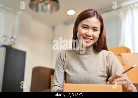 Schöne junge asiatische weibliche E-Commerce-Unternehmer bereitet ihr Lieferpaket, Schreiben einer Kundenadresse auf Karton. KMU online Stockfoto