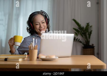 Glückliche ältere asiatische Frau, die Kopfhörer trägt, hört im Wohnzimmer des gemütlichen Heims Lieder auf dem Laptop Stockfoto