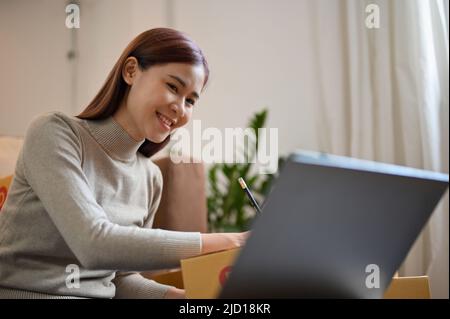Wunderschöne, tausendjährige asiatische Frau, die auf den Laptop-Bildschirm schaut und die Adresse ihres Kunden auf einen Versandkarton schreibt. Kleinunternehmen, Online-Unternehmen, e-comm Stockfoto