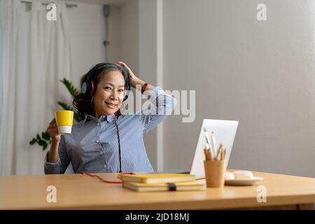 Glückliche ältere asiatische Frau, die Kopfhörer trägt, hört im Wohnzimmer des gemütlichen Heims Lieder auf dem Laptop und schaut auf die Kamera. Stockfoto