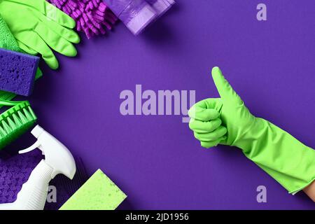 Weibliche Hand mit Daumen nach oben im Schutzhandschuh und Reinigungsprodukte auf violettem Hintergrund. Housekeeping- und Hausarbeit-Konzept. Flach liegend, Draufsicht. Stockfoto