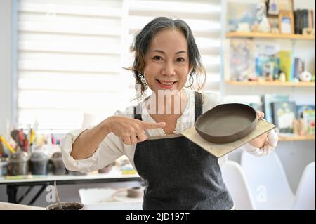 Professionelle gealterte asiatische Töpferin zeigt ihre Keramikplatte in ihrem Studio. Schöne gealterte asiatische Frau, die sich einer Töpferwerkstatt anschließt. Stockfoto