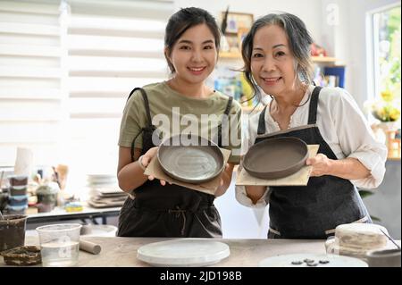 Porträt einer schönen asiatischen jungen Frau mit einer attraktiven asiatisch gealterten Frau, die eine handgefertigte Schale und einen Teller in der Handwerkswerkstatt hält. Stockfoto