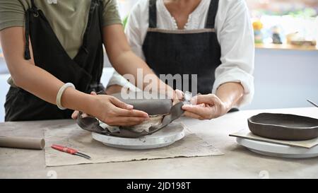 Professionelle gealterte Lehrerin, die einer schönen jungen Frau beibringt, in der Werkstatt eine Keramikschale mit Ton zu machen. Zugeschnittenes Bild Stockfoto