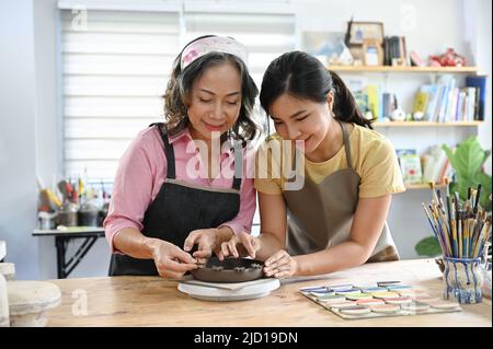 Freundliche junge asiatische Handwerkerin, die einer älteren Frau beibringt, im Atelier eine Tonerde zu machen. Stockfoto