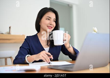 Eine attraktive junge asiatische Geschäftsfrau oder Geschäftsführerin sitzt an ihrem Schreibtisch und hat einen heißen Kaffee. Stockfoto