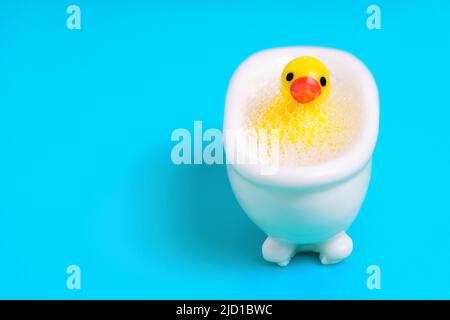 Kleine gelbe Ente in einer Spielzeugbadewanne voller Seifenblasen isoliert auf blauem Hintergrund mit Kopierraum. Hygienegewohnheiten für Kinder. Stockfoto