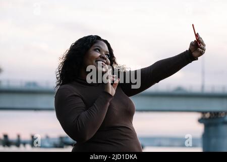 Lockige afroamerikanische pralle Frau im Kleid, die Selfie macht, Entengesicht per Smartphone macht und sich vor dem Sonnenuntergang posiert Stockfoto