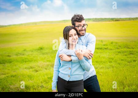 Porträt eines glücklichen Paares, das sich auf dem Feld umarmt, Porträt eines jungen Paares, das im grünen Feld verliebt ist und die Kamera anschaut, Porträt eines niedlichen Paares Stockfoto